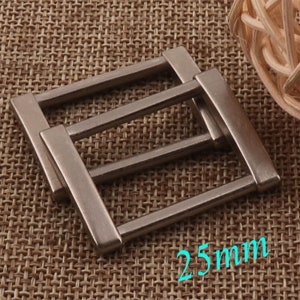 May include: Two silver metal belt buckle slides, each measuring 25 mm in length. The slides are rectangular with three horizontal bars.
