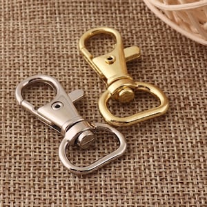 May include: Two metal swivel clasps, one silver and one gold, on a brown woven fabric background.