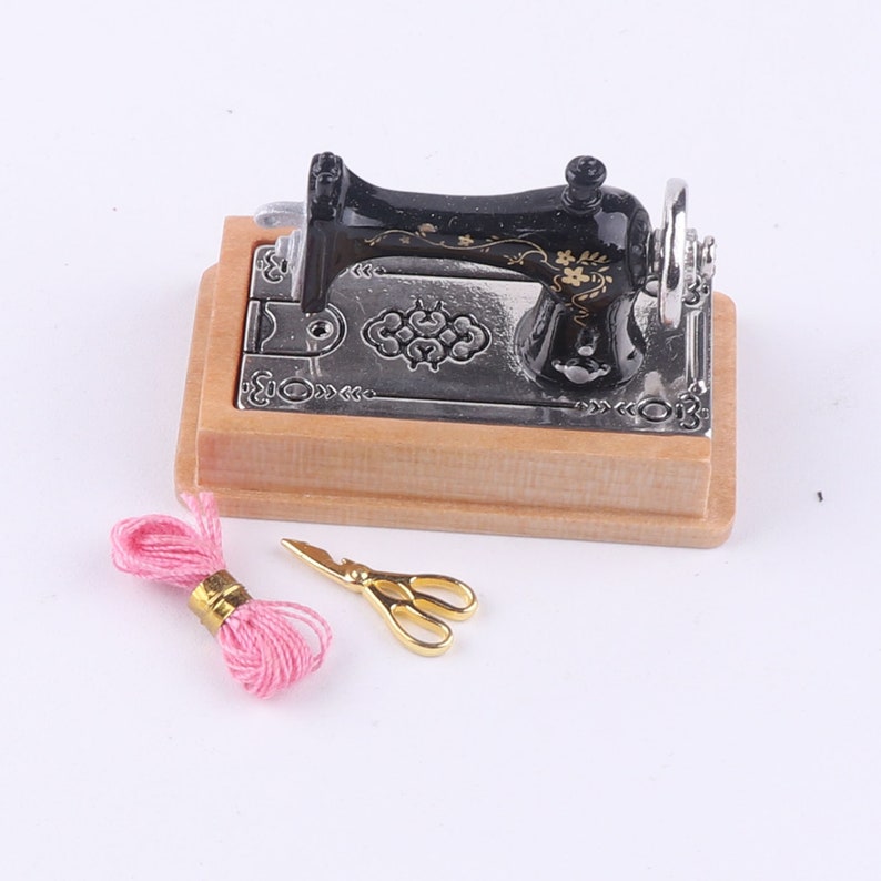 Może przedstawiać: Miniaturowa czarna maszyna do szycia z drewnianą podstawą, r&oacute;żową nicią i złotymi nożyczkami.