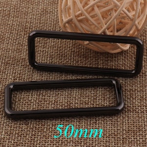 May include: Two black metal rectangular rings, 50mm in size, on a brown woven fabric background.