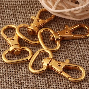 May include: Four gold-colored metal clasps with a hook and a loop. The clasps are on a brown woven surface.