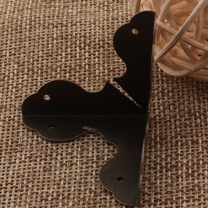 May include: Two black metal corner brackets with a decorative design. The brackets are on a brown burlap background.