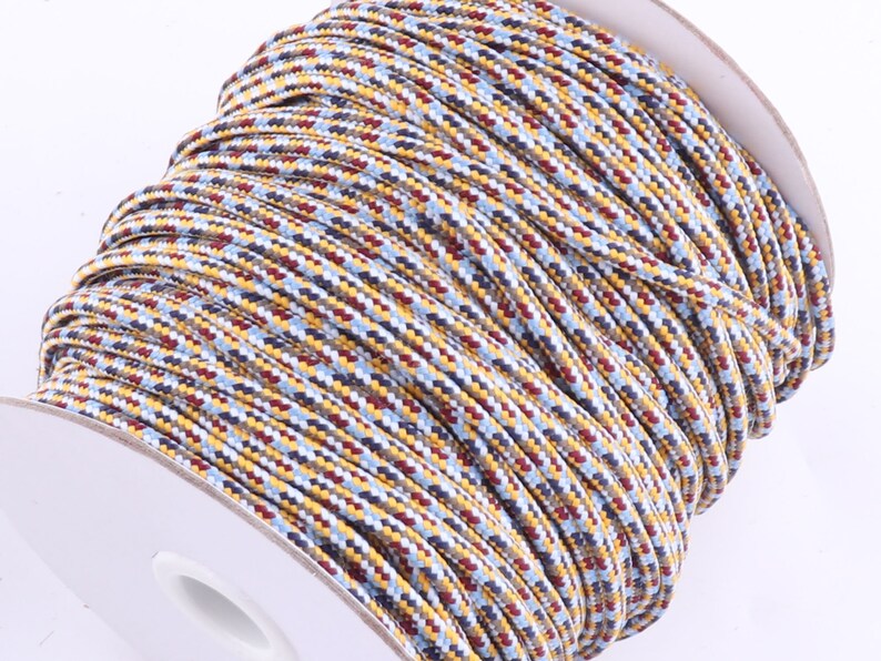 Op de afbeelding: Een spoel met veelkleurige koord op een witte achtergrond. Het koord is gemaakt van verschillende kleuren, waaronder rood, blauw, geel en bruin.