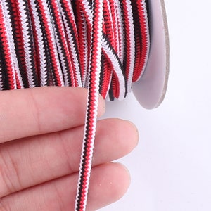 May include: Red, black, and white striped elastic cord. The cord is wound around a spool and a hand is holding a section of the cord.