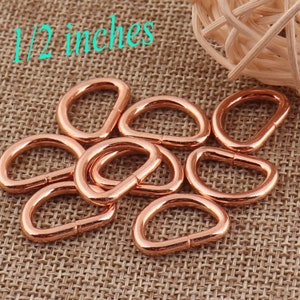 May include: A close-up of ten rose gold D-rings, each measuring 1/2 inch in diameter. The rings are arranged in a pile on a brown burlap fabric.