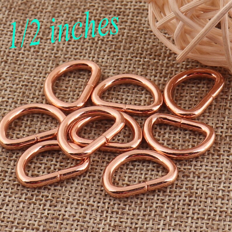 May include: A close-up of ten rose gold D-rings, each measuring 1/2 inch in diameter. The rings are arranged in a pile on a brown burlap fabric.