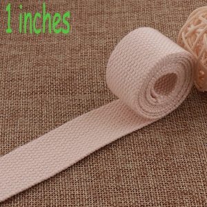 以下が含まれることがあります： 麻の生地の上に置かれた、幅2.5cmのピンク色の綿のウェビングストラップのクローズアップ。ストラップは、質感のあるパターンで織られています。