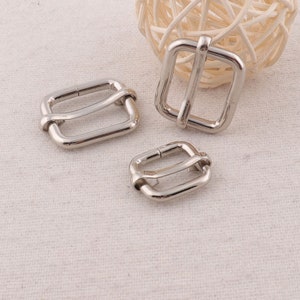 May include: Three silver metal slider buckles of different sizes. The buckles are rectangular with a rounded edge and have a single, centred, horizontal bar.