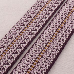 Puede incluir: Dos cintas tejidas de color morado y blanco con detalles de hilo dorado. La cinta tiene un patrón geométrico.
