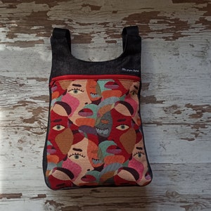 May include: A tote bag featuring a vibrant abstract face design. The bag has black straps and a red zip. The bag is made of woven fabric and has the text "Jela popa fuma" on the top.