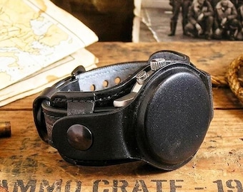 May include: A black leather watch case with a silver watch face, resting on a wooden ammo crate. The case has a button closure and a light blue interior. The crate is stamped with the words "AMMO CRATE - 194" and a star.