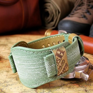 May include: A green canvas watch strap with a tan leather backing and accents. The strap is on top of a watch with a silver case. The text "DURE. DISCOVER." is at the top of the image.