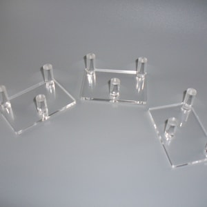May include: Three clear acrylic display stands, each with four small cylindrical feet. The stands are rectangular and have a smooth, polished surface.