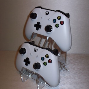 May include: Two white Xbox controllers are displayed on a clear acrylic stand. The controllers have black buttons and a black Xbox logo.