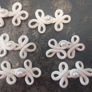 Può includere: Sei chiusure a nodo cinese in tessuto bianco con un accento di perla al centro di ogni nodo.