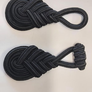 Könnte beinhalten: Zwei schwarze Seil-Knöpfe mit einem geknoteten Design. Die Knöpfe haben eine Schlaufe an einem Ende und ein abgerundetes Ende am anderen.