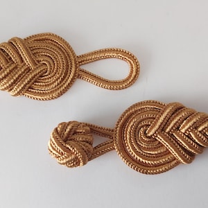 May include: Two gold-colored decorative rope toggles. The toggles are made of braided rope and have a circular shape with a loop on one end. The toggles are a decorative element for clothing or home decor.