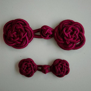 Peut inclure: Deux ensembles de boutons chinois en tissu bordeaux. L'ensemble le plus grand comporte deux gros boutons et un bouton de connexion plus petit. L'ensemble le plus petit comporte deux boutons plus petits et un bouton de connexion plus petit.