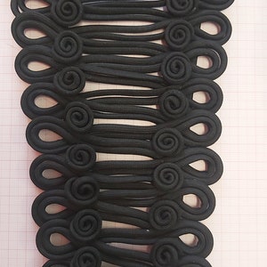 May include: A set of 19 black fabric toggle buttons with a looped design. The buttons are arranged in a vertical row on a gridded surface.