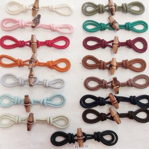 Könnte beinhalten: Ein Set mit 14 Paaren Kordelverschlüssen mit Holzknöpfen. Die Kordelverschlüsse sind in verschiedenen Farben erhältlich, darunter Weiß, Rosa, Rot, Orange, Grau, Hellblau, Beige, Grün, Türkis, Rotbraun, Braun, Schwarz und Dunkelbraun.