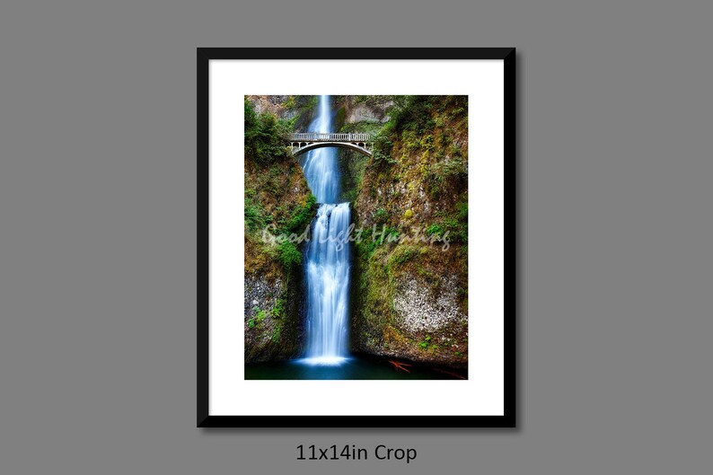 May include: A photograph of a waterfall cascading down a rocky cliff face. A wooden bridge spans the top of the waterfall, with lush green foliage surrounding the water.