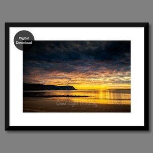 May include: A scenic photograph of a sunrise over a body of water with a dark, cloudy sky. The sun is reflecting off the water, creating a golden glow. The horizon is marked by a distant shoreline and a small hill.