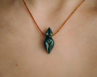 May include: A dark green ceramic pendant shaped like a woman's body, hanging from a brown leather cord.