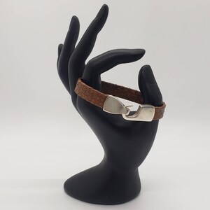 May include: A brown leather bracelet with a silver clasp. The bracelet is on a black hand-shaped display stand.