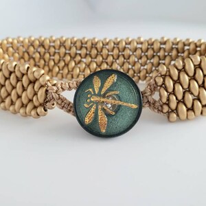 May include: Gold-toned beaded bracelet with a central dark green button clasp. The button features a gold dragonfly design with a small crystal accent. The bracelet is made of small, oval-shaped beads.