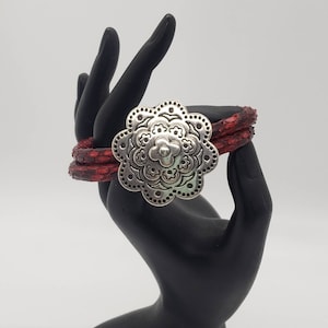 May include: A red leather bracelet with a silver floral design. The bracelet is on a black hand model.