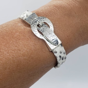 May include: A silver buckle bracelet with a white leather strap. The buckle has a textured surface and a rounded shape.