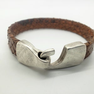 May include: A brown leather bracelet with a silver clasp. The bracelet is made of braided leather and has a unique, textured design.