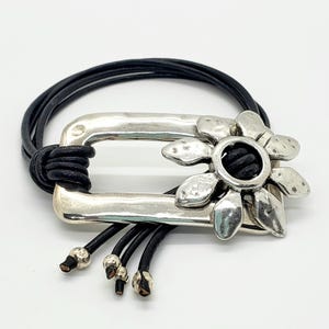 May include: A silver-toned metal bracelet with a rectangular center and a flower-shaped accent. The bracelet features black leather cords and a decorative tassel. The flower has a black center and the bracelet is on a white background.