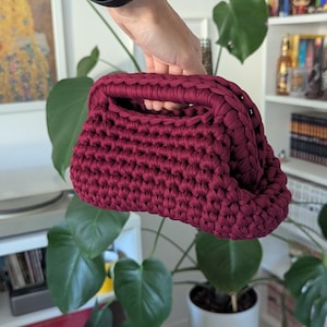 Op de afbeelding: Een bordeauxrode gehaakte clutch met een afgeronde vorm en een handvat. De tas is gemaakt van dik garen, met zichtbare steken die een textuur creëren. De clutch wordt omhooggehouden tegen een achtergrond van groene planten en een witte boekenplank.