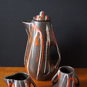 May include: A brown ceramic teapot and two matching creamer cups with white and orange abstract designs.