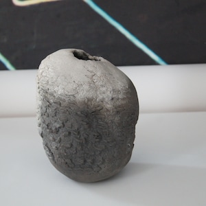 May include: A grey ceramic vase with a rough, textured surface. The vase has a small opening at the top.