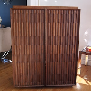 May include: A mid-century modern wooden cabinet with two doors. The doors feature a unique, textured design with vertical grooves. The cabinet is a rich brown colour and has a sleek, minimalist design.
