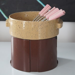 Peut inclure: Un service à fondue vintage avec un pot en céramique beige moucheté et une base marron foncé. Le pot a deux poignées et contient des fourchettes à fondue avec des manches roses. Un article de cuisine rétro.