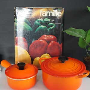Peut inclure: Deux casseroles en émail orange avec des couvercles et des poignées noirs sont présentées devant un livre de cuisine intitulé "famille" avec une illustration colorée de légumes. Les casseroles sont sur une surface blanche avec un fond sombre.