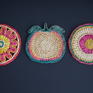 May include: Three woven pot holders in a circular shape. The first pot holder is a bright pink, yellow, and green flower design. The second pot holder is a green, pink, and yellow tomato shape. The third pot holder is a pink, yellow, and green concentric circle design.