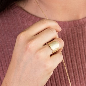 Könnte beinhalten: Ein goldener Hammerring mit einem breiten Band und einer abgerundeten Oberseite. Der Ring wird am Zeigefinger einer Hand getragen.