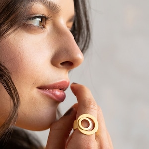 May include: A gold ring with two concentric circles. The ring is worn on the index finger of a person's hand.