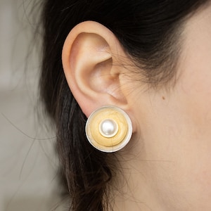 May include: A pair of silver and gold earrings with a pearl in the center. The earrings are round and have a concentric circle design.