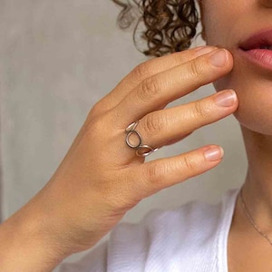 May include: A silver ring featuring a design of interlocked circles. The ring is worn on the index finger of a person's hand.