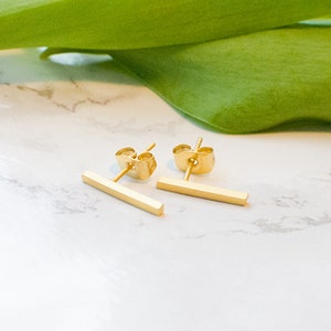 Könnte beinhalten: Ein Paar goldene Bar-Ohrstecker. Die Ohrringe sind schlicht und minimalistisch, mit einem geraden Barren-Design.