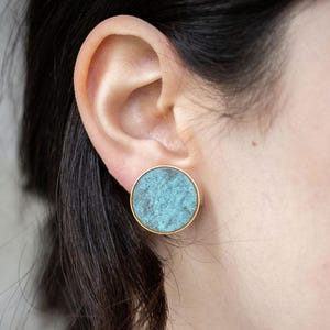 以下が含まれることがあります： 金色の縁取りのある丸いターコイズブルーのイヤリングのペア。イヤリングは、テクスチャのある大理石のような外観をしています。