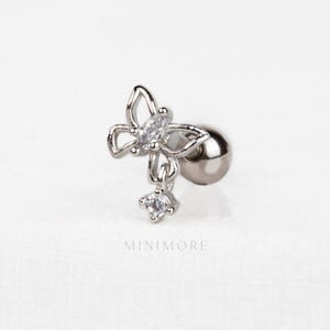 May include: A silver earring featuring a butterfly design with a dangling gemstone. The butterfly has clear gemstones in the wings. The earring has a ball-shaped backing. The word "MINIMORE" is printed at the bottom.