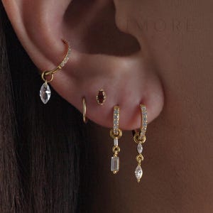 May include: A collection of gold-toned hoop earrings featuring clear crystal charms in marquise and baguette shapes. The set includes a small plain hoop, a dark red stone stud, and multiple pavé-set hoops with dangling crystal pendants.
