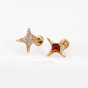 Könnte beinhalten: Zwei goldfarbene Ohrstecker mit einem vierzackigen Sterndesign. Ein Ohrstecker ist mit einem klaren, quadratisch geschliffenen Stein besetzt, der andere mit einem orangefarbenen quadratischen Stein. Darunter steht das Wort MINIMORE in hellgrau.