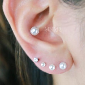 Op de afbeelding: Een set van vier oorpiercings met één witte bolvormige parel in de oorschelp en drie knopjes in de oorlel, bestaande uit één kleine heldere ronde edelsteen en twee witte parels van verschillende groottes, met de tekst MNMR zichtbaar.